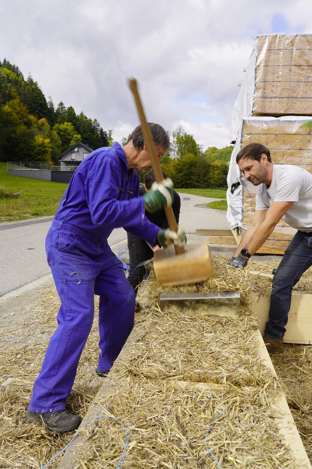 Zwei Personen verdichten mit einem Holzschlägel Strohballen in einem hölzernen Wandelement auf der Baustelle