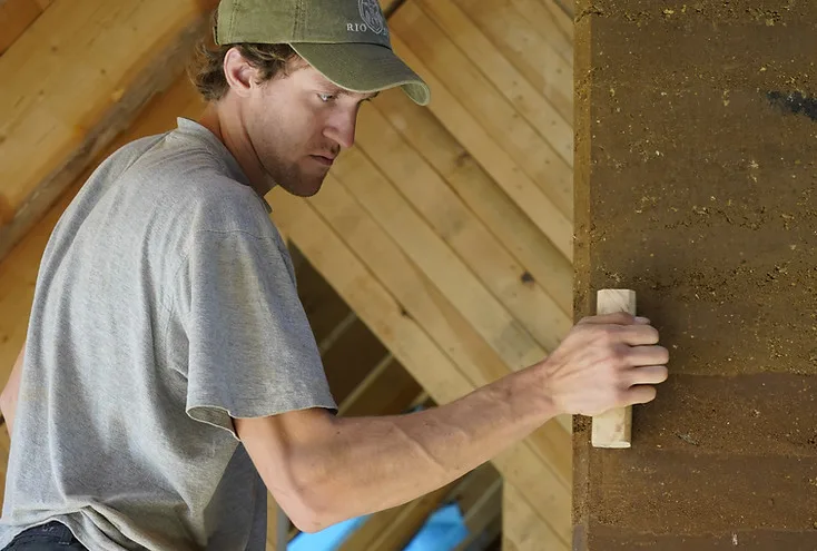 Handwerker glättet mit einem Holzwerkzeug die Oberfläche einer Stampflehmwand unter einem hölzernen Dachstuhl