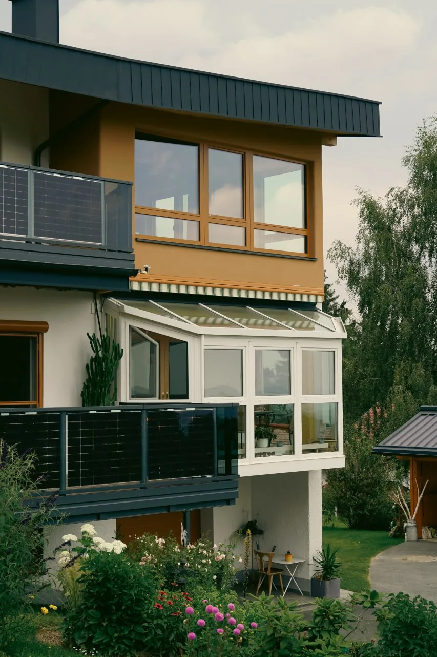 Haus Anita: Detail der Südseite mit Photovoltaikmodulen am Balkongeländer und Wintergarten im bestehenden Geschoss