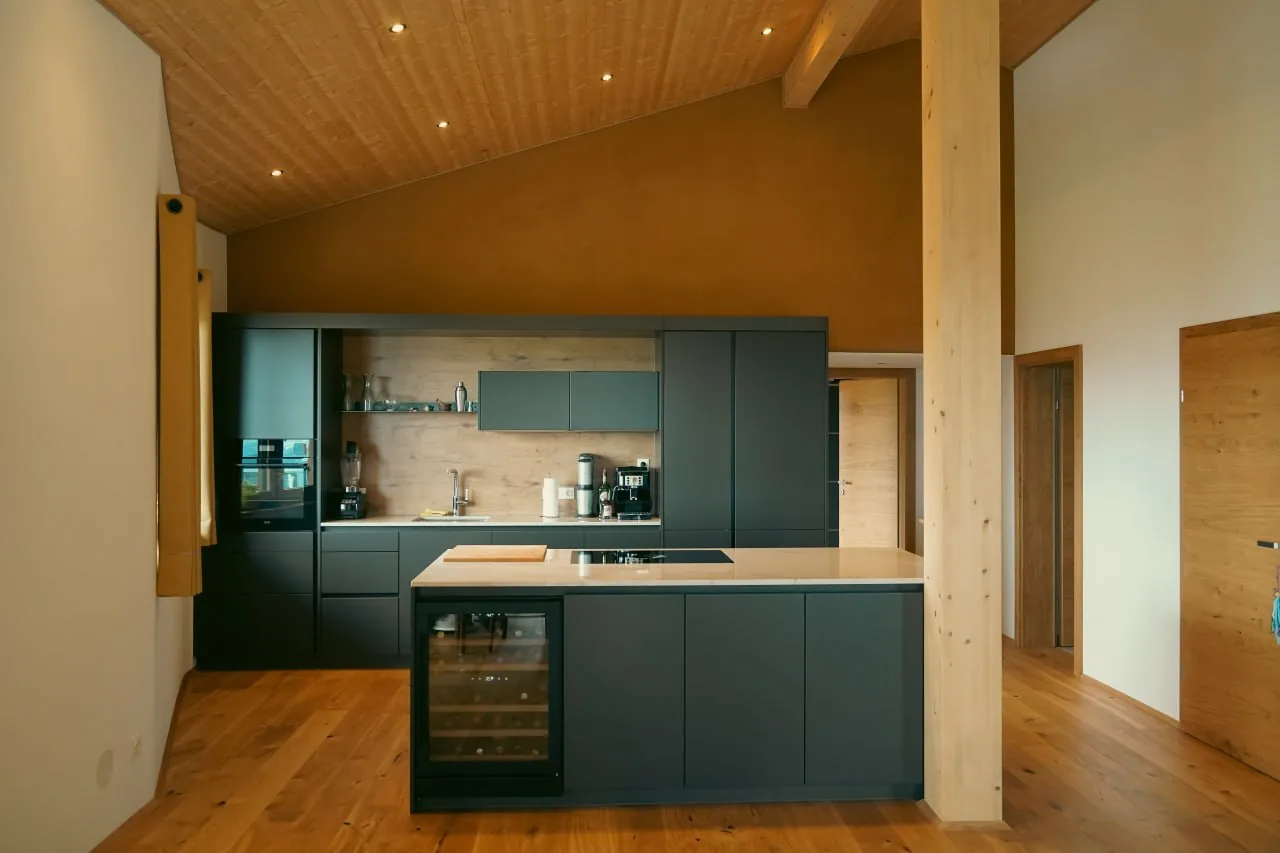 Haus Anita: Küche mit dunkelgrünen Fronten vor einer Lehmputzwand, darüber eine Holzdecke bis zum First