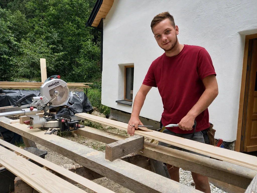 Zimmerer Max beim Zuschnitt von Kanthölzern auf einer Baustelle, im Hintergrund ein weiß verputztes Haus